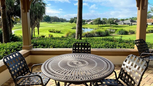 A covered patio with a round metal table and four chairs, overlooking a golf course and a lake.