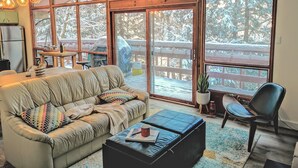 A cozy living room with a sofa, a coffee table, and a chair. There is a large window with a view of a snowy deck outside. - 3 BR Harp Hygge House. Only 8min to Hidden Valley! (Huntsville)