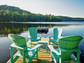 Une jetée en bois, ornée de chaises Adirondack vertes, fait face à un lac paisible, le tout étant entouré d’arbres et d’une colline lointaine.