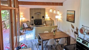 A cozy living area with a fireplace, wooden ceiling beams, a dining table with chairs, and a kitchen area with a stove. - 3 BR Harp Hygge House. Only 8min to Hidden Valley! (Huntsville)