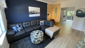 Ein modernes Wohnzimmer mit dunkelblauer Akzentwand, einem schwarzen Ledersofa im Eckdesign, einem gemusterten Hocker und einem Deckenventilator.