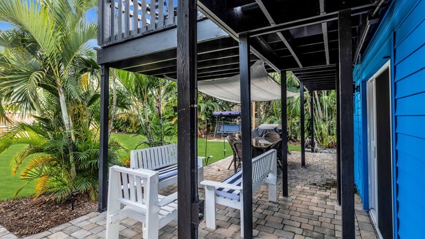 Eine überdachte Terrasse mit weißen Bänken, einem Grill und Blick auf einen Garten mit Palmen.