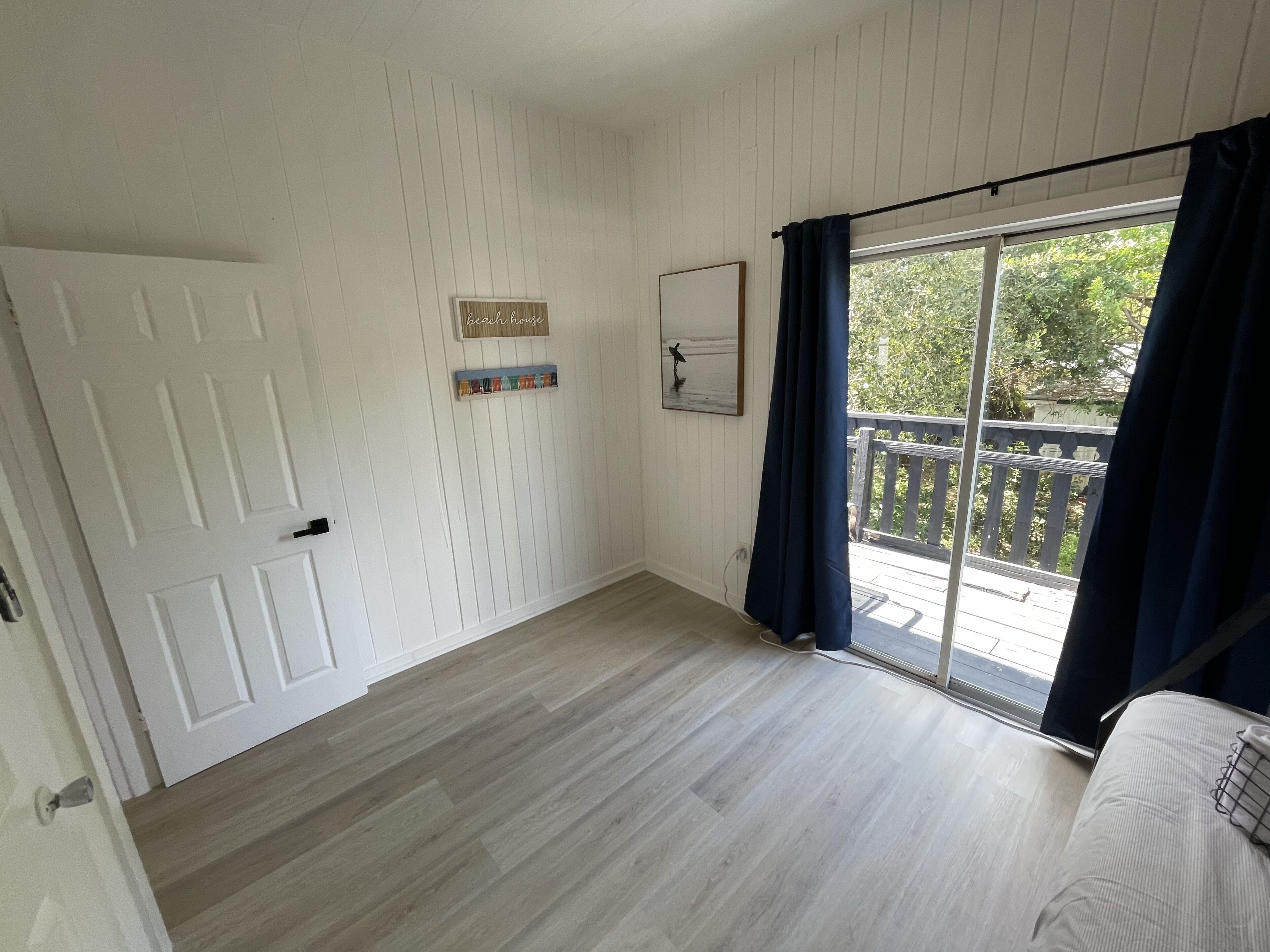 Een kamer met een houten vloer, een witte deur, een raam met donkere gordijnen en uitzicht op een houten terras.