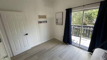 Een kamer met een houten vloer, een witte deur, een raam met donkere gordijnen en uitzicht op een houten terras.