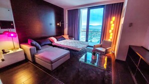 Ein modernes Hotelzimmer mit einem Bett, einer Couch, einem kleinen Tisch und einem Sessel. Ein großes Fenster bietet Blick auf die Stadt.