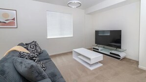 A modern living room with a grey sofa, a flat-screen TV on a glass stand, a white coffee table, and a ceiling light fixture.