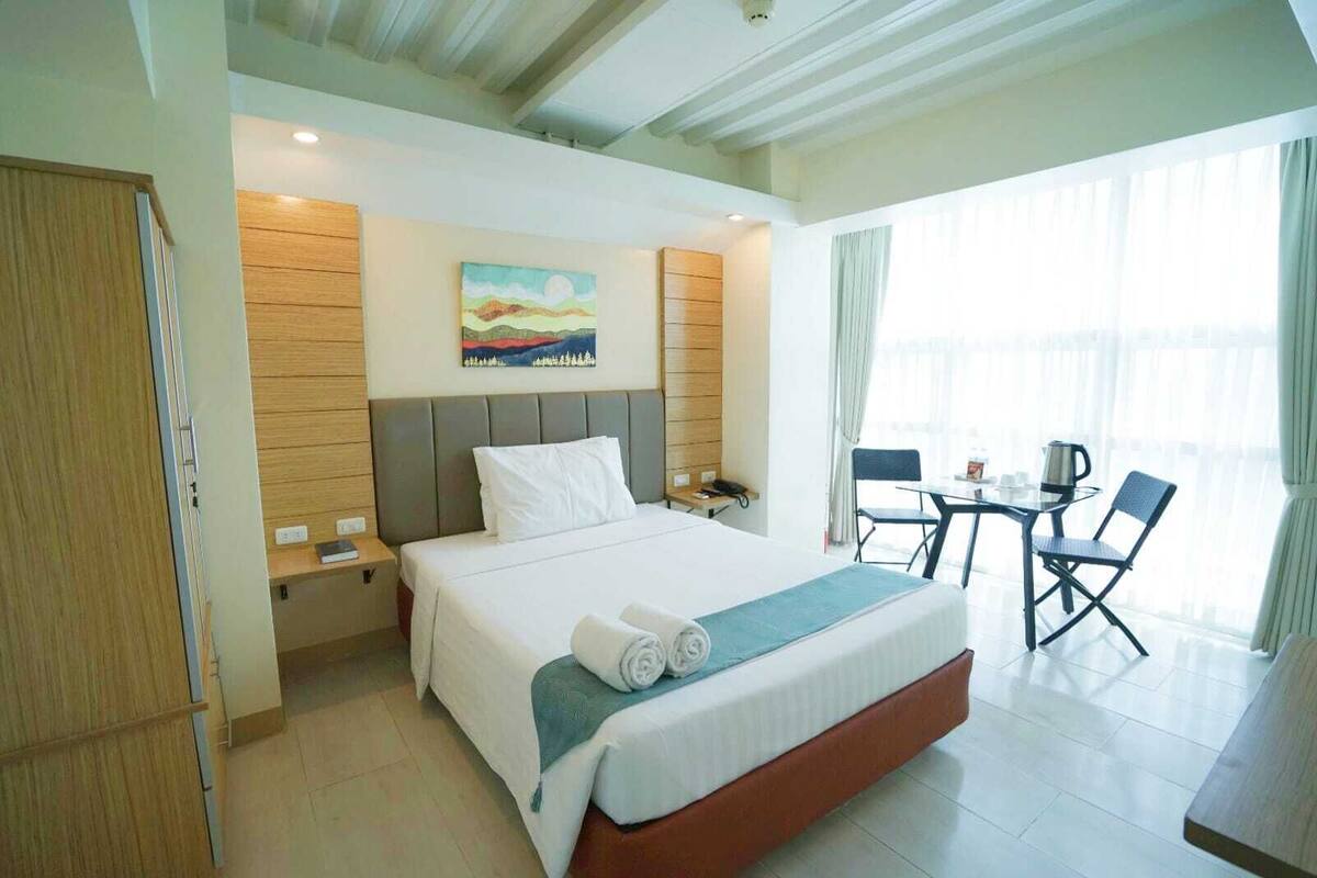 Una habitación de hotel con una cama, una mesita de noche, un cuadro en la pared, un escritorio con una silla y una ventana con cortinas.