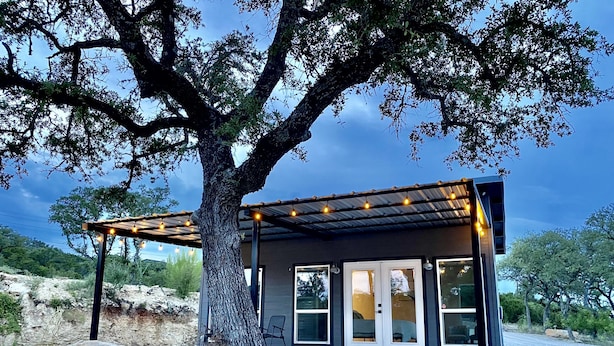 Une maison moderne dotée d’une pergola et de guirlandes lumineuses, entourée d’une allée en gravier et d’un grand arbre.