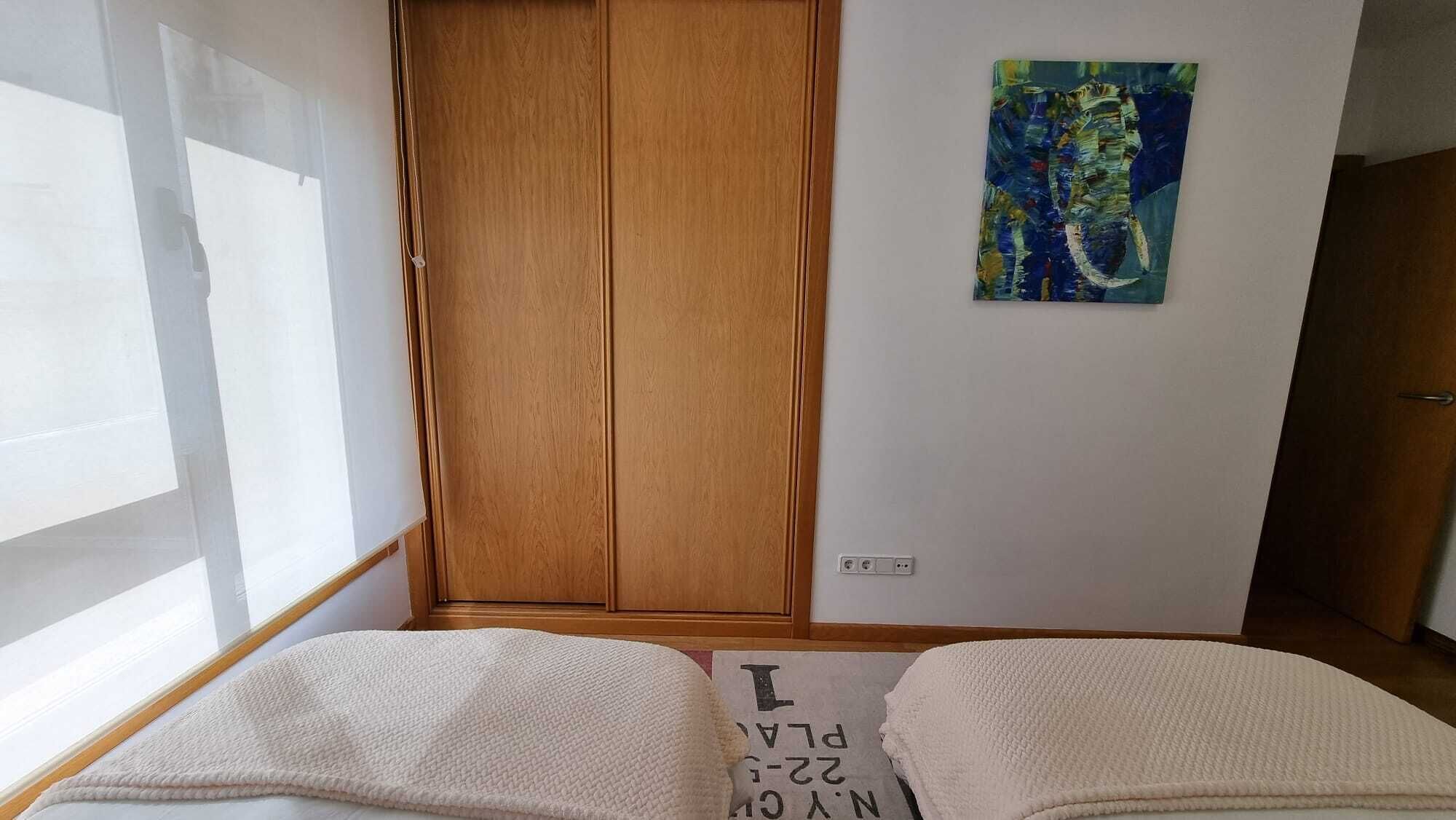 Une chambre avec une armoire en bois, un tableau accroché au mur et un lit recouvert d’une couverture blanche.