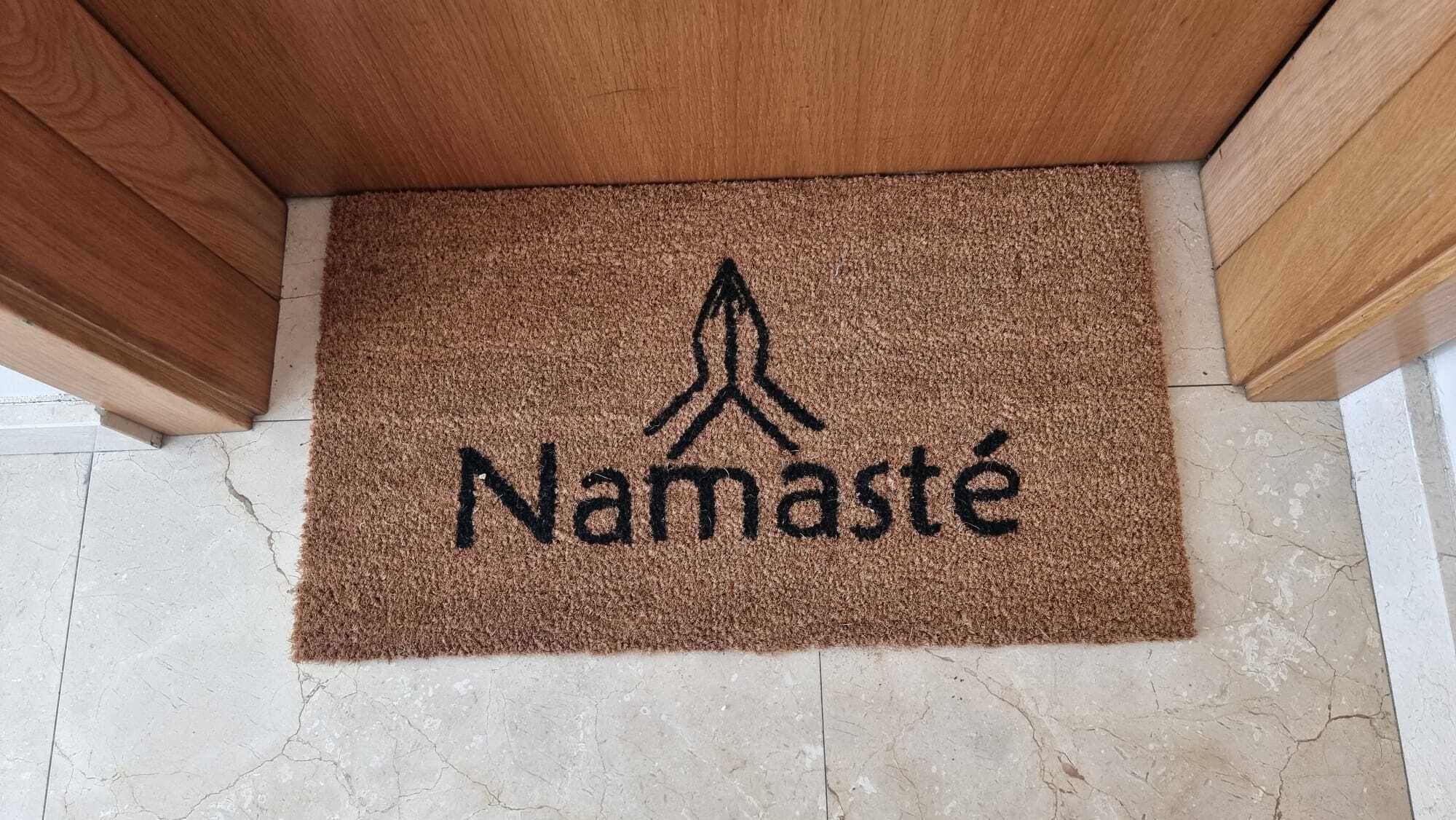 Un paillasson marron orné d’une empreinte de main et du mot « Namaste », posé sur un sol carrelé.