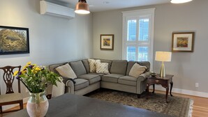 A living room with a gray sectional sofa, a coffee table, and a vase of flowers. There are two pendant lights, a window with blinds, and framed artwork on the walls.