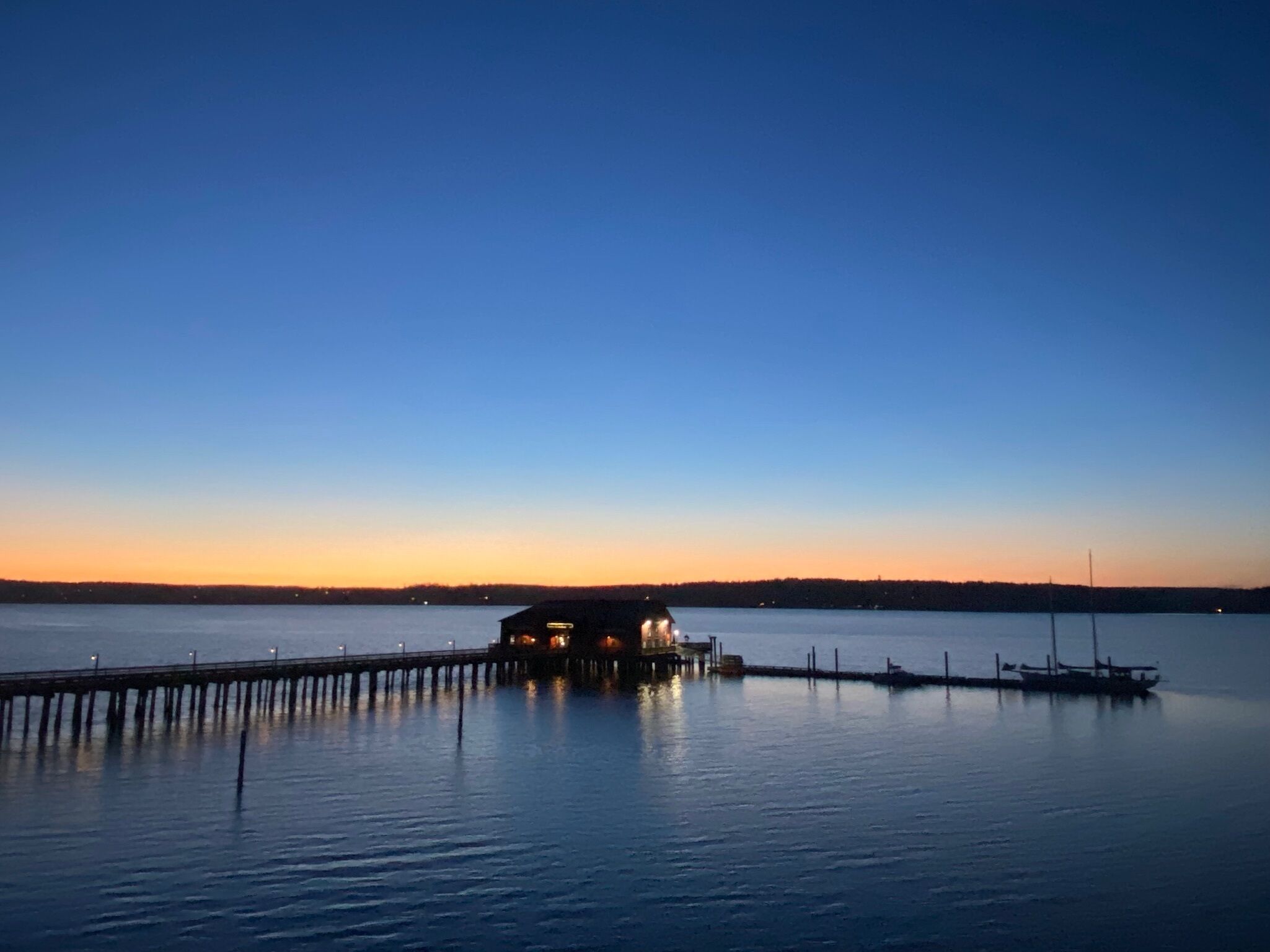 A waterfront property with a dock extending into calm water, a building with lights on, and a clear sky at dusk.