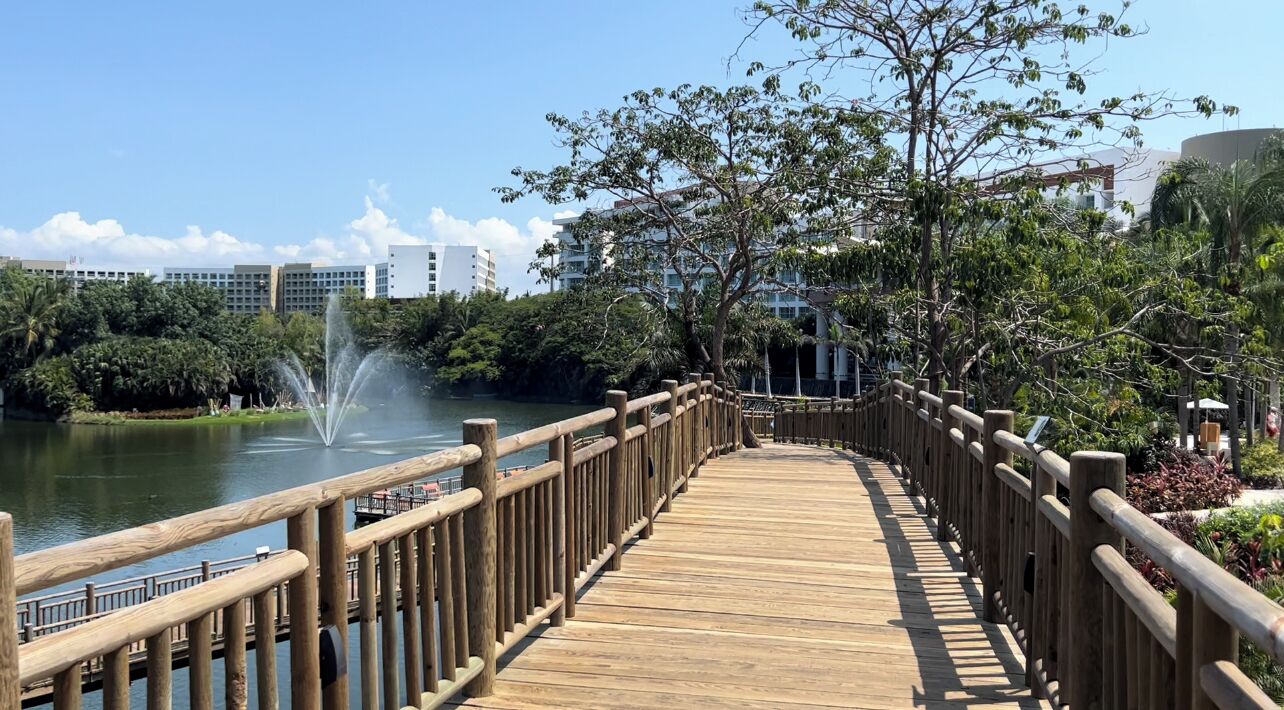 Um calçadão de madeira com uma fonte, cercado por vegetação e edifícios ao fundo.