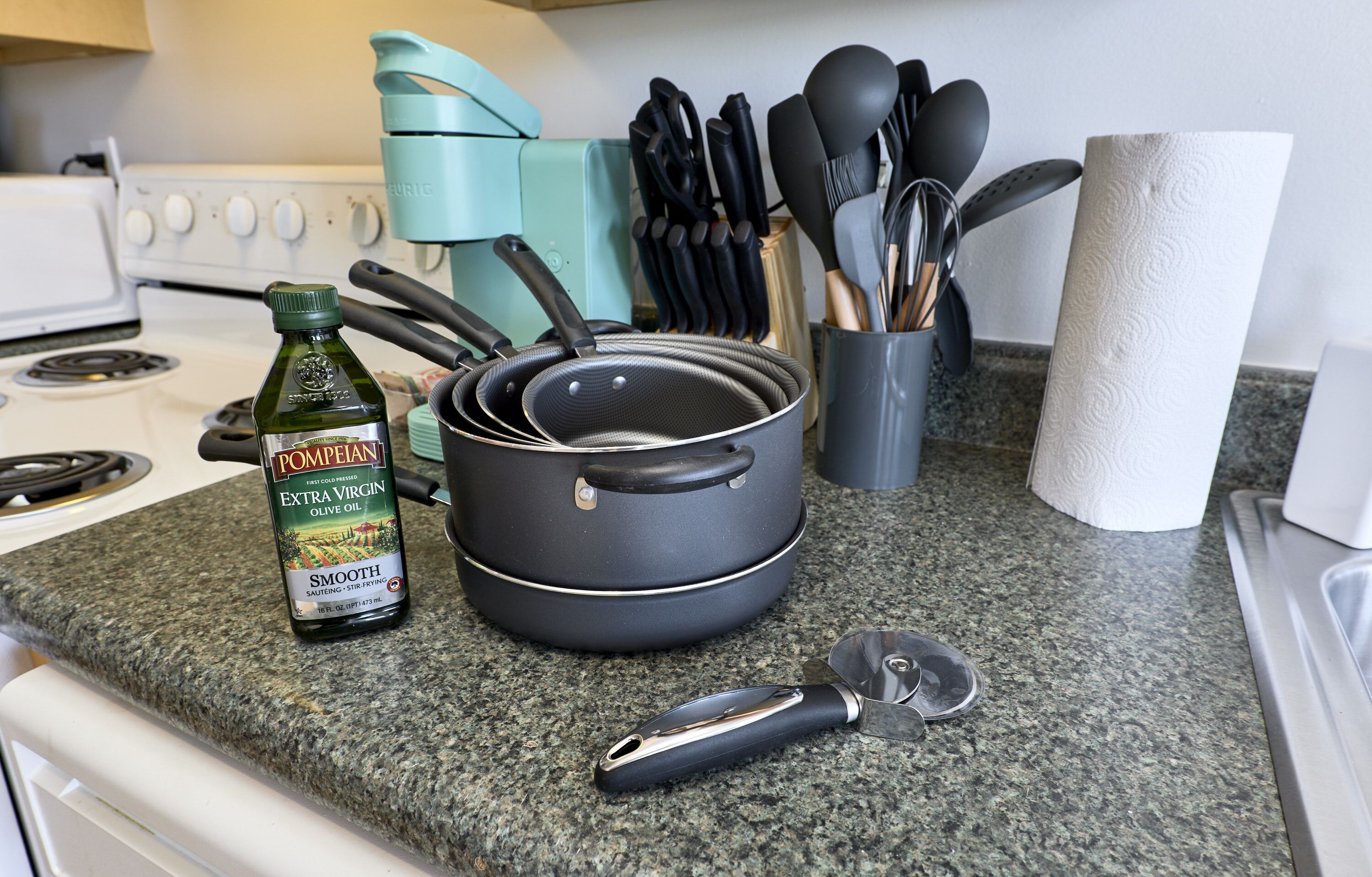 Un piano di lavoro in cucina con una bottiglia di olio extravergine d'oliva, una pentola nera, una grattugia e diversi utensili da cucina.