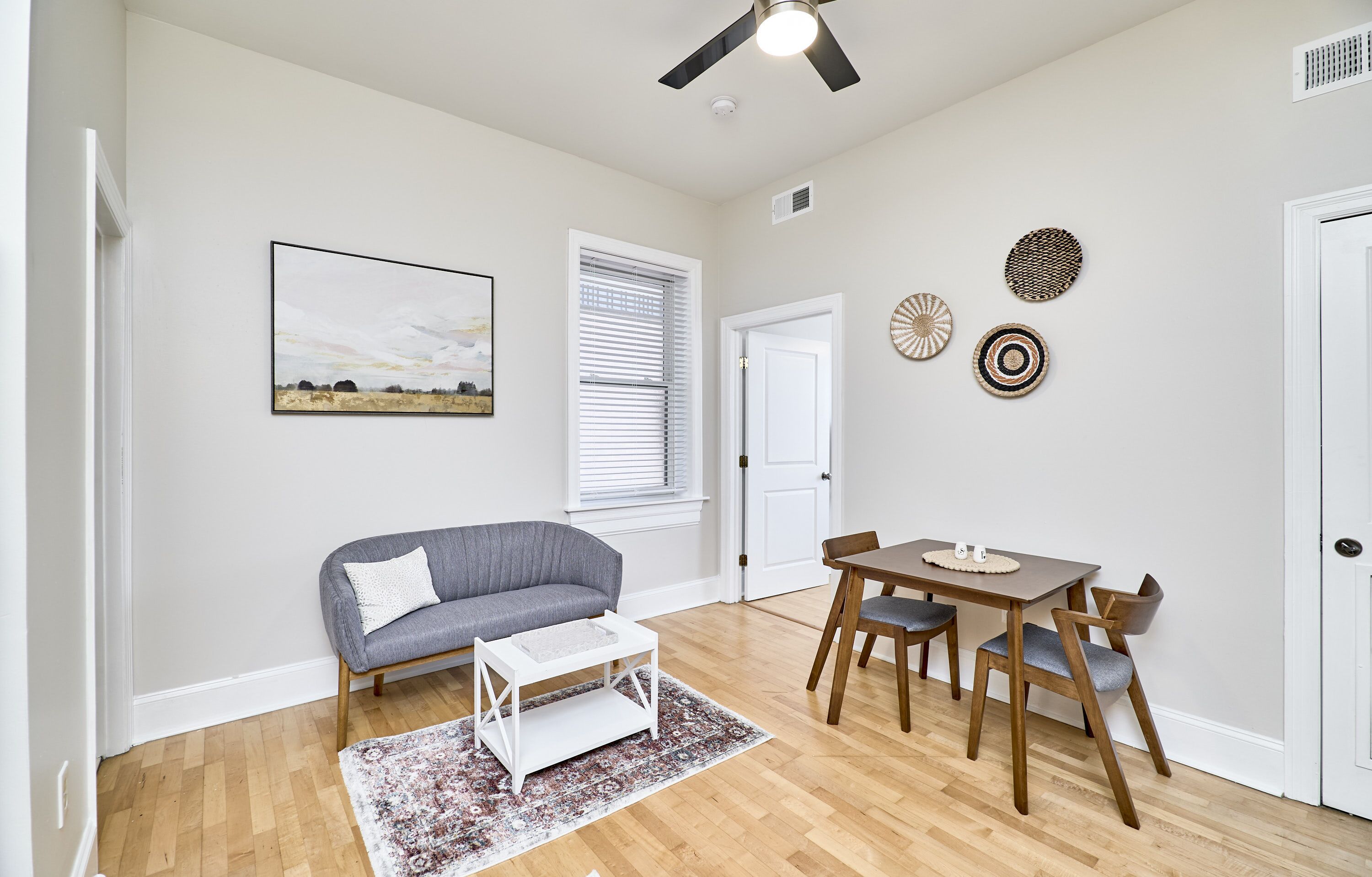 A living room with a gray sofa, a white coffee table, a wooden dining table with chairs, a ceiling fan, and a painting on the wall.