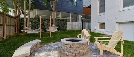 Un patio trasero con un hogar de piedra, dos sillas Adirondack y una hamaca.