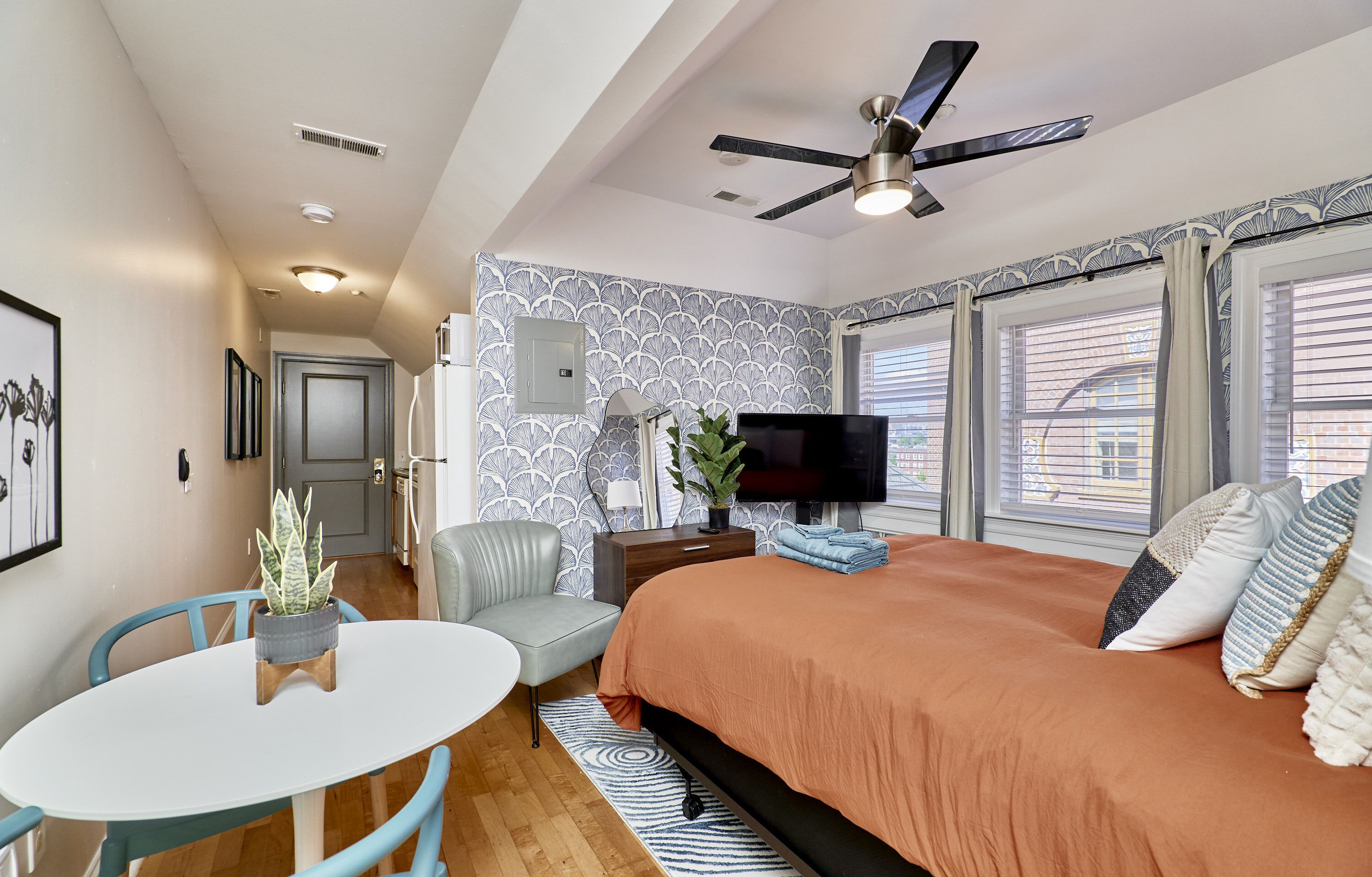A bedroom with a bed, a TV, a ceiling fan, a plant, a chair, and a table.