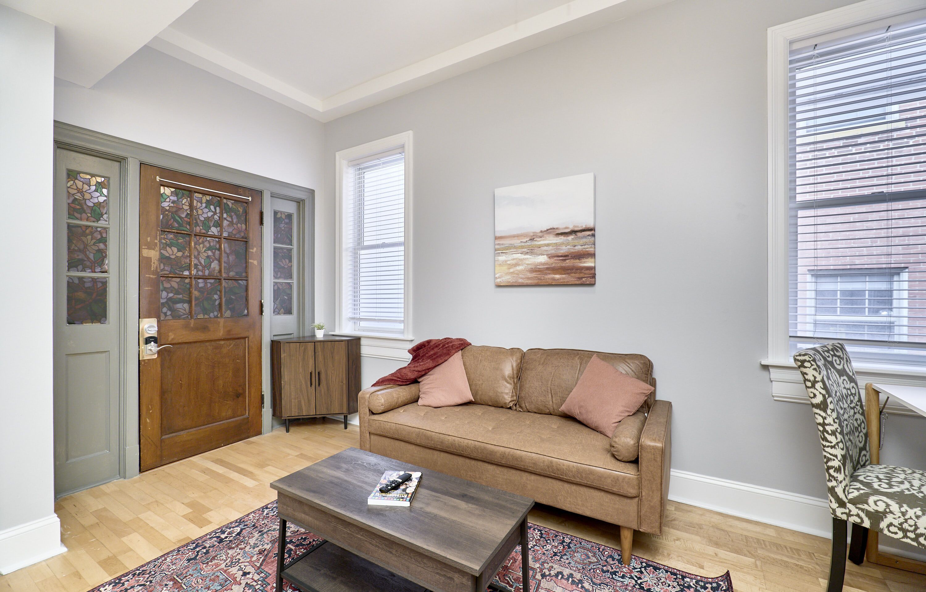 A living room with a brown leather sofa, a wooden coffee table, a patterned rug, and a window with blinds.
