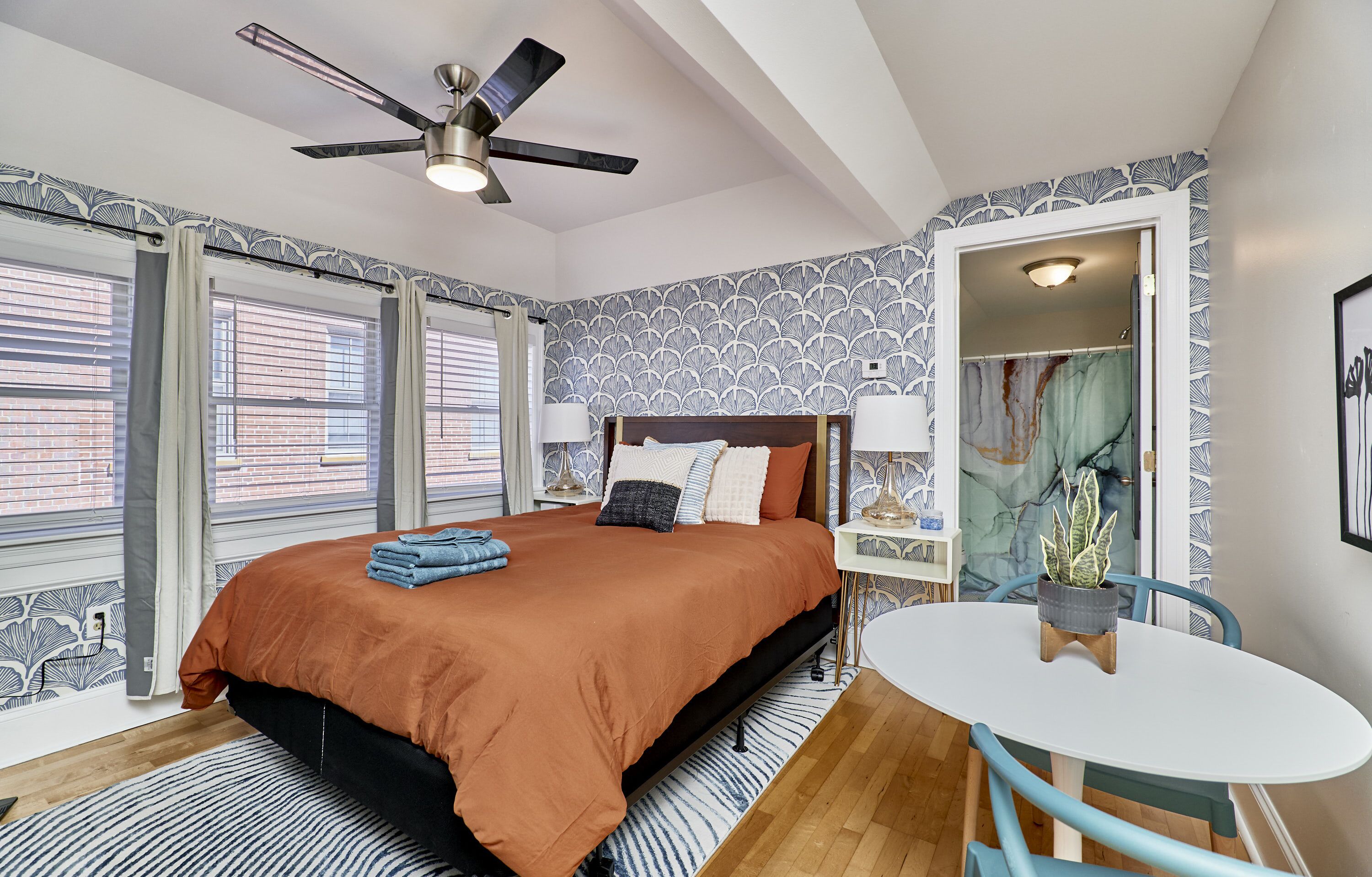 A bedroom with a bed, a ceiling fan, a window with blinds, a nightstand, a table with a chair, and a potted plant.