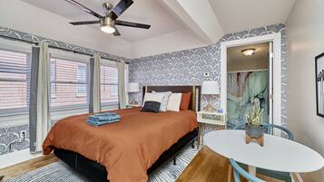 A bedroom with a bed, a ceiling fan, a window with blinds, a nightstand, a table with a chair, and a potted plant.
