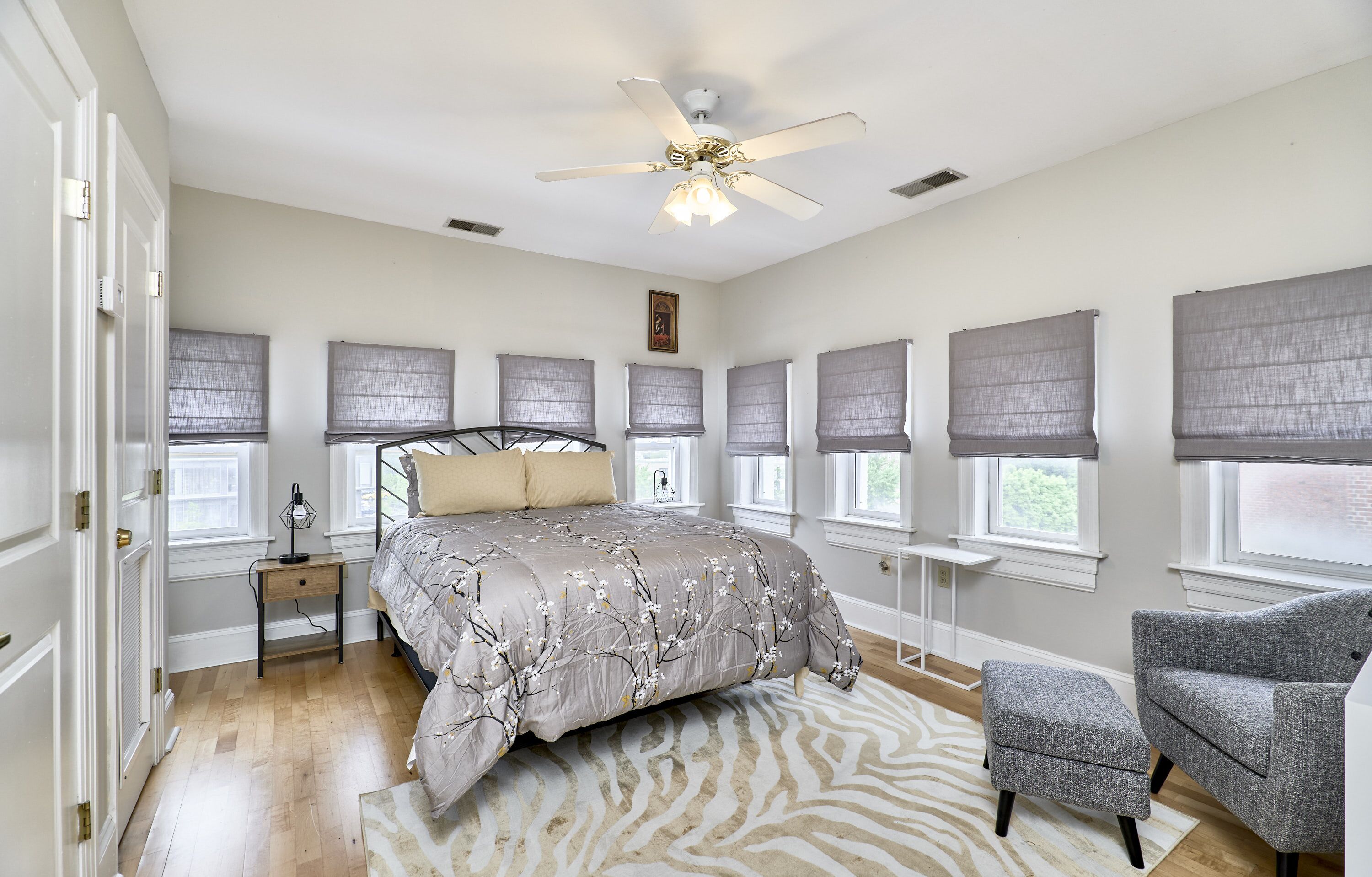 A bedroom with a bed, a chair, a nightstand, a ceiling fan, and multiple windows with blinds.