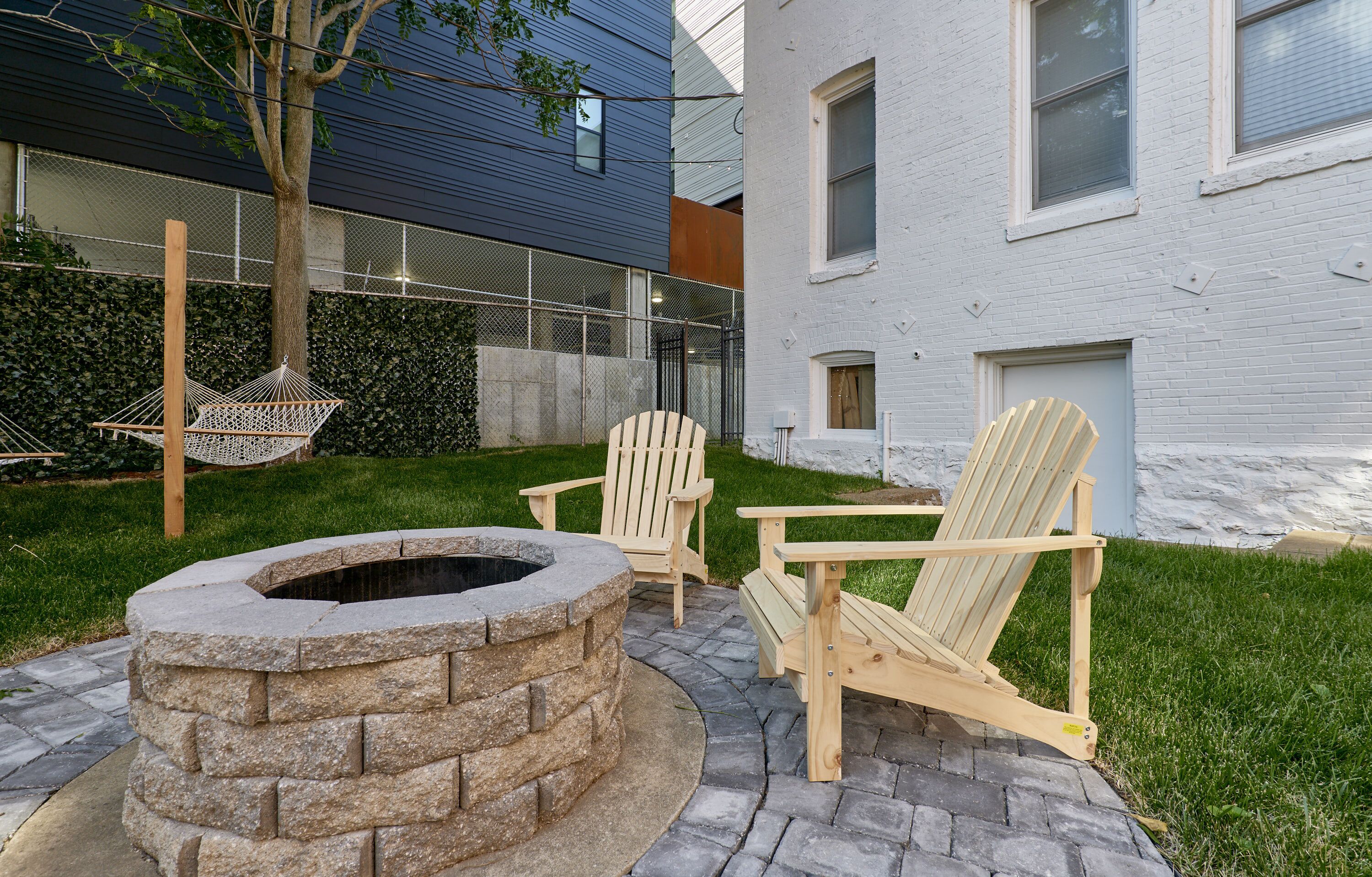 A stone fire pit with two Adirondack chairs and a hammock in a landscaped outdoor area.