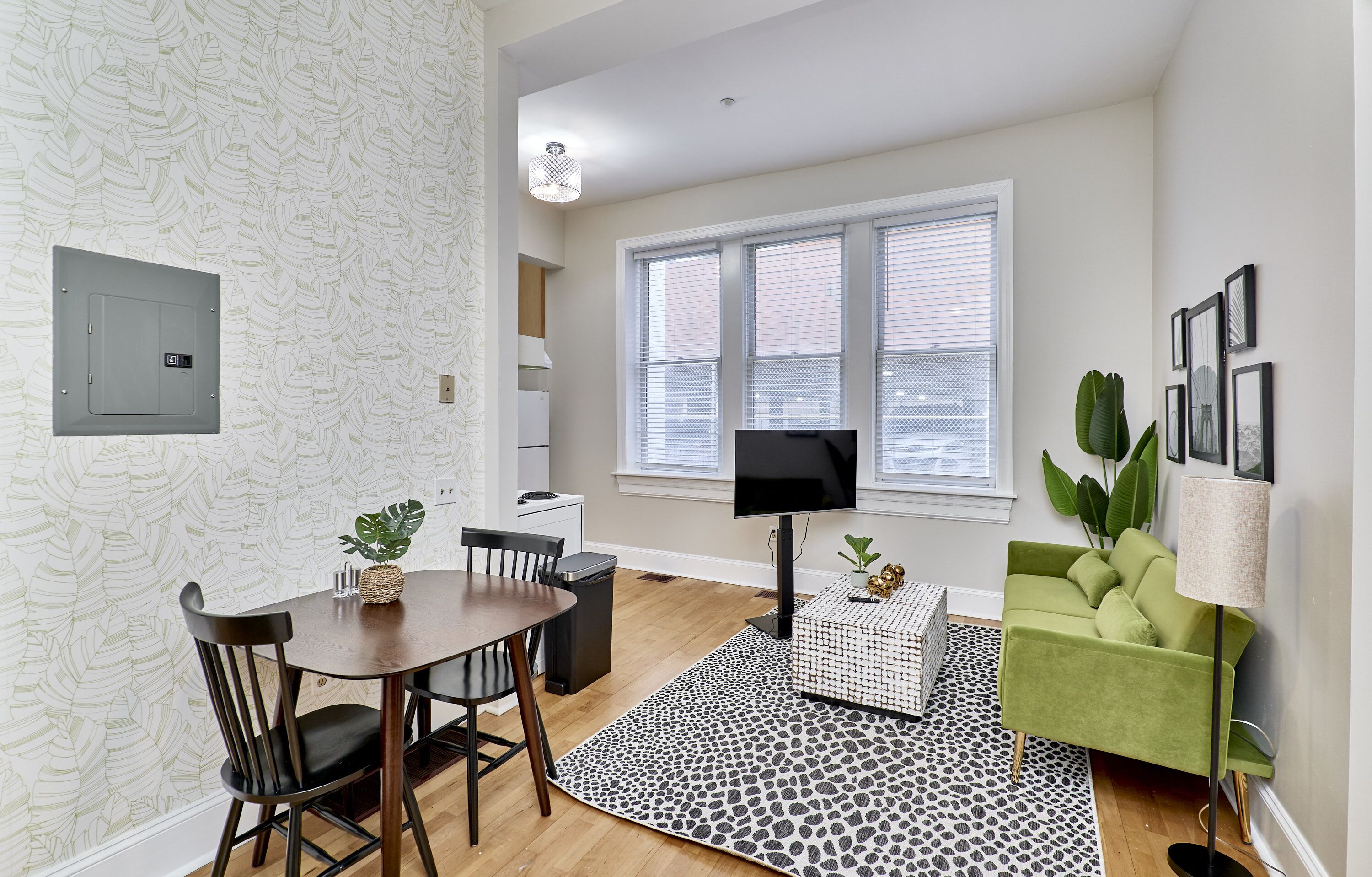 A modern living room with a dining area, featuring a large window, a flat-screen TV, a green sofa, and a patterned rug.