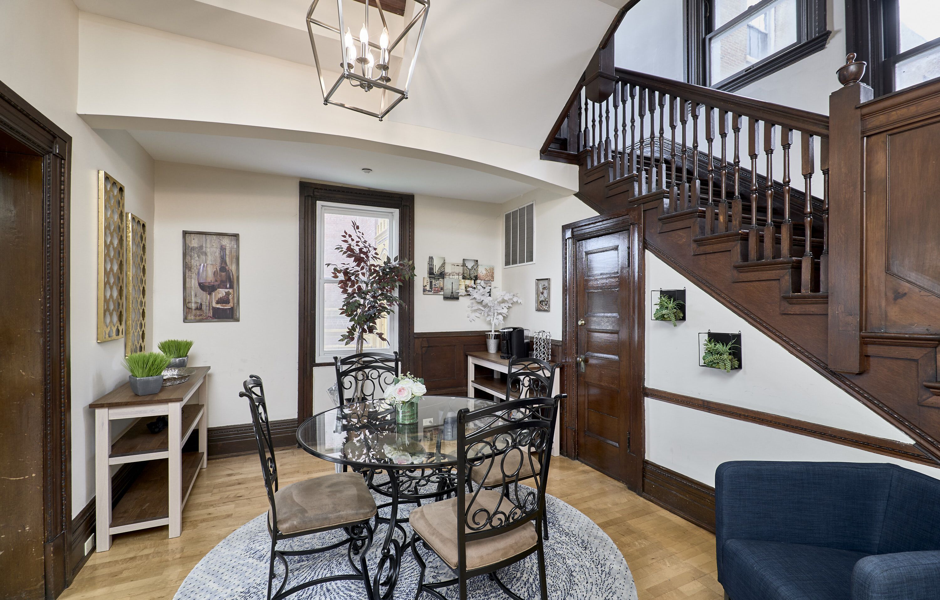 A spacious living area with a round glass table, dark wooden chairs, and a blue sofa. A wooden staircase with a railing leads to an upper floor.