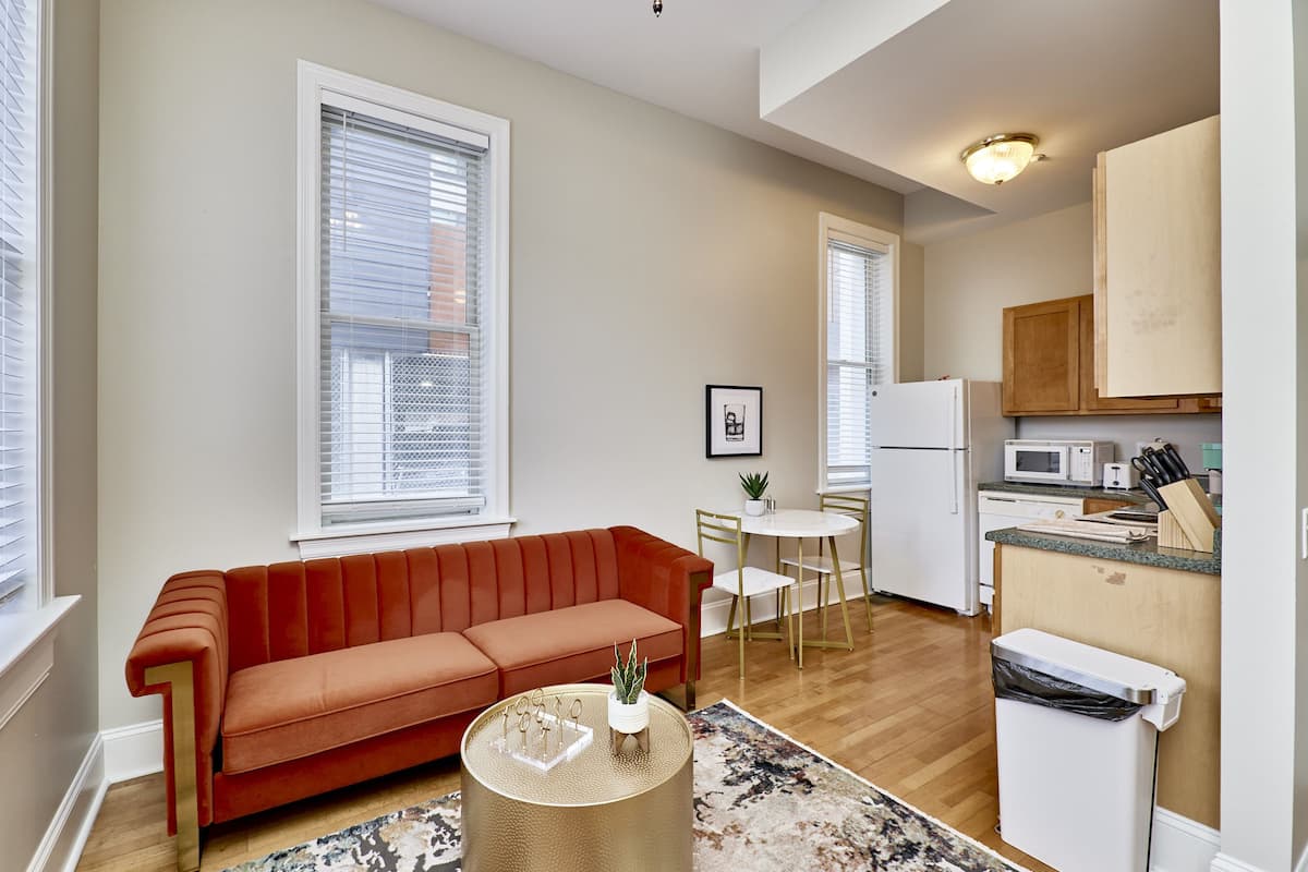 A compact living space with a sofa, a dining table, a kitchen area, and a trash bin.