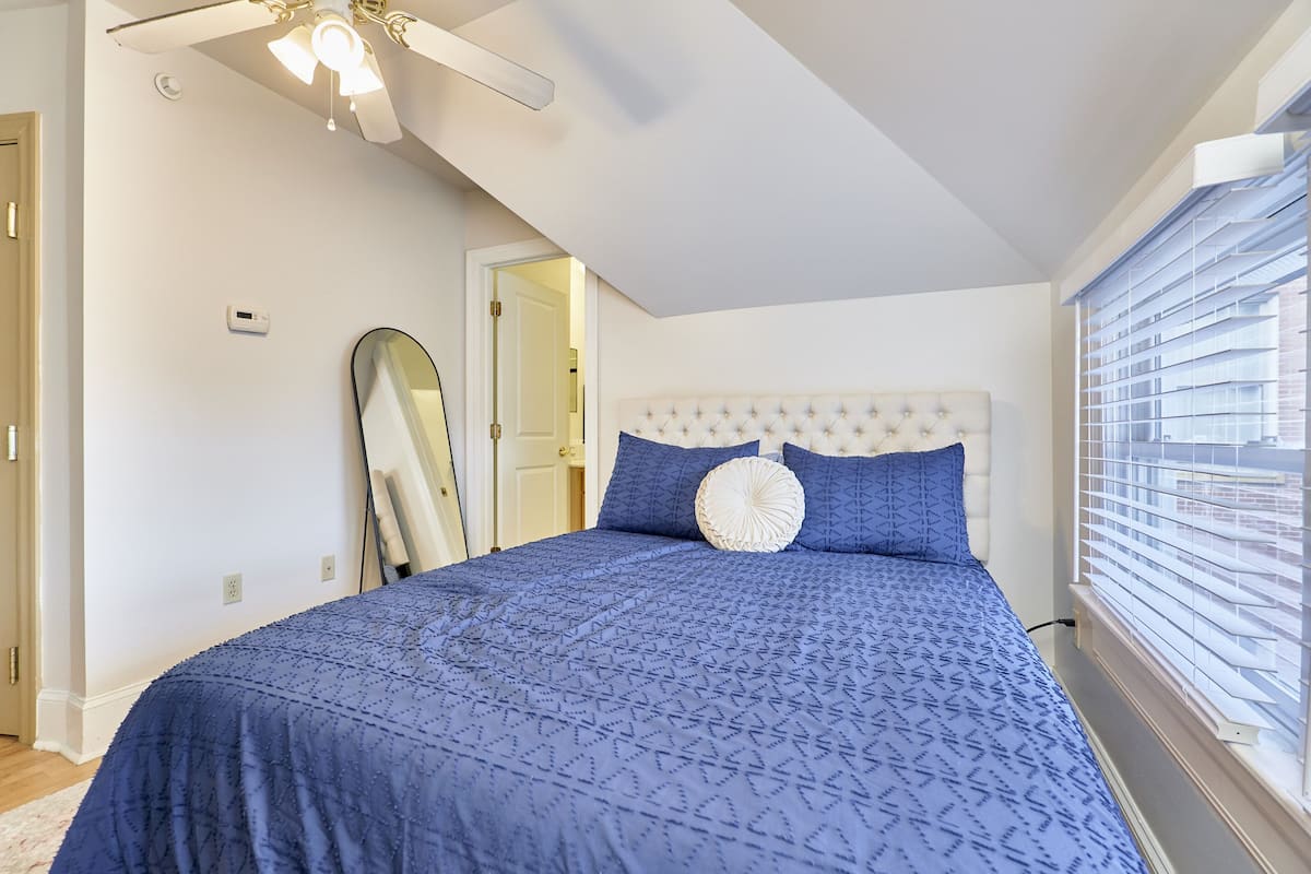 A bedroom with a large bed, a ceiling fan, a window with blinds, and a door.