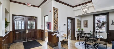 A spacious lobby with a wooden floor, a reception desk, a 'WELCOME' sign, a dining area with a table and chairs, and a large window.