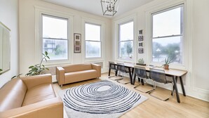 A spacious living room with a beige sofa, a dining area with a table and chairs, large windows, and a chandelier. -  White House at Grand Central (St. Louis)