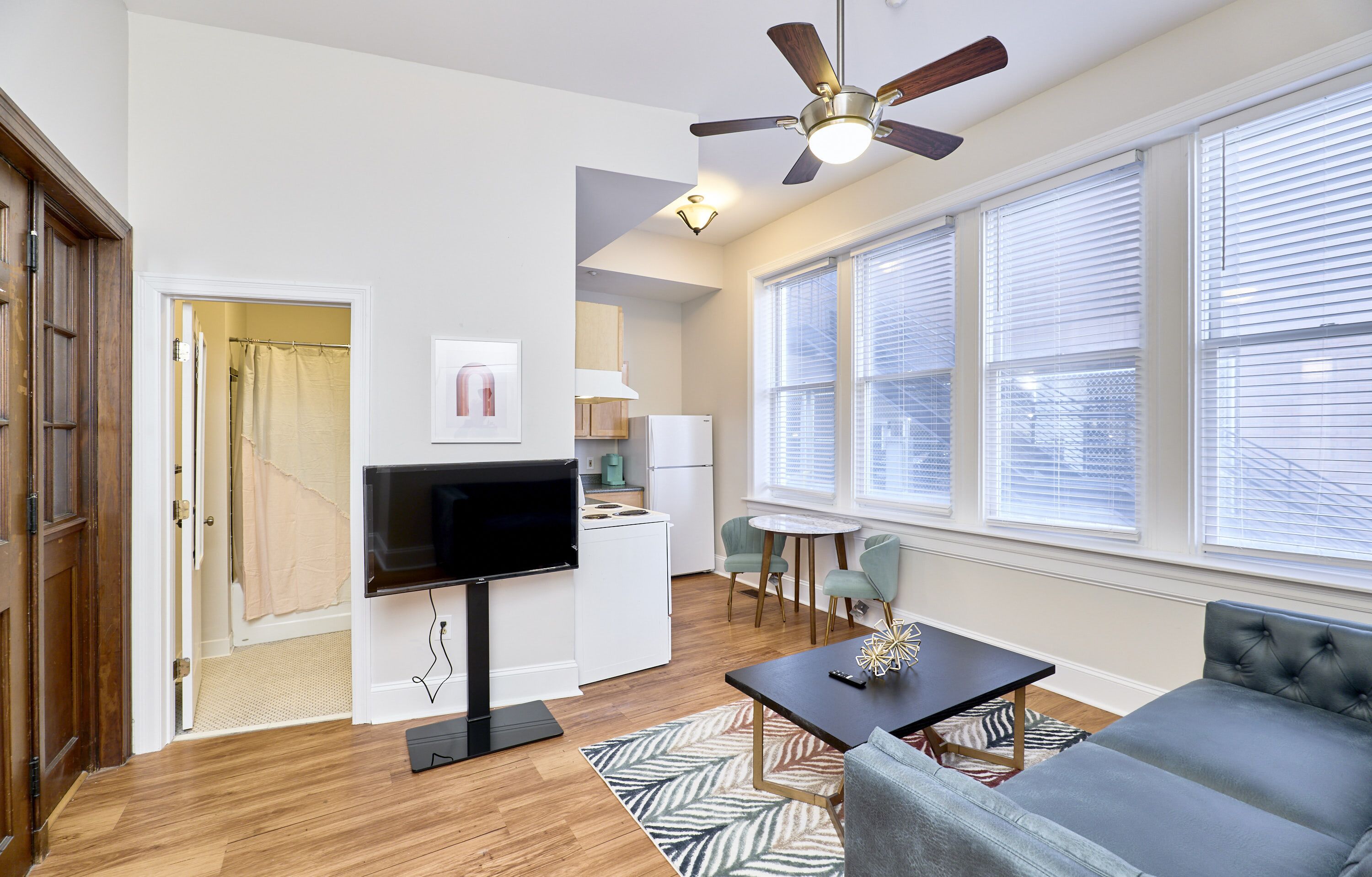 A modern living room with a flat-screen TV, a ceiling fan, and a large window with blinds.