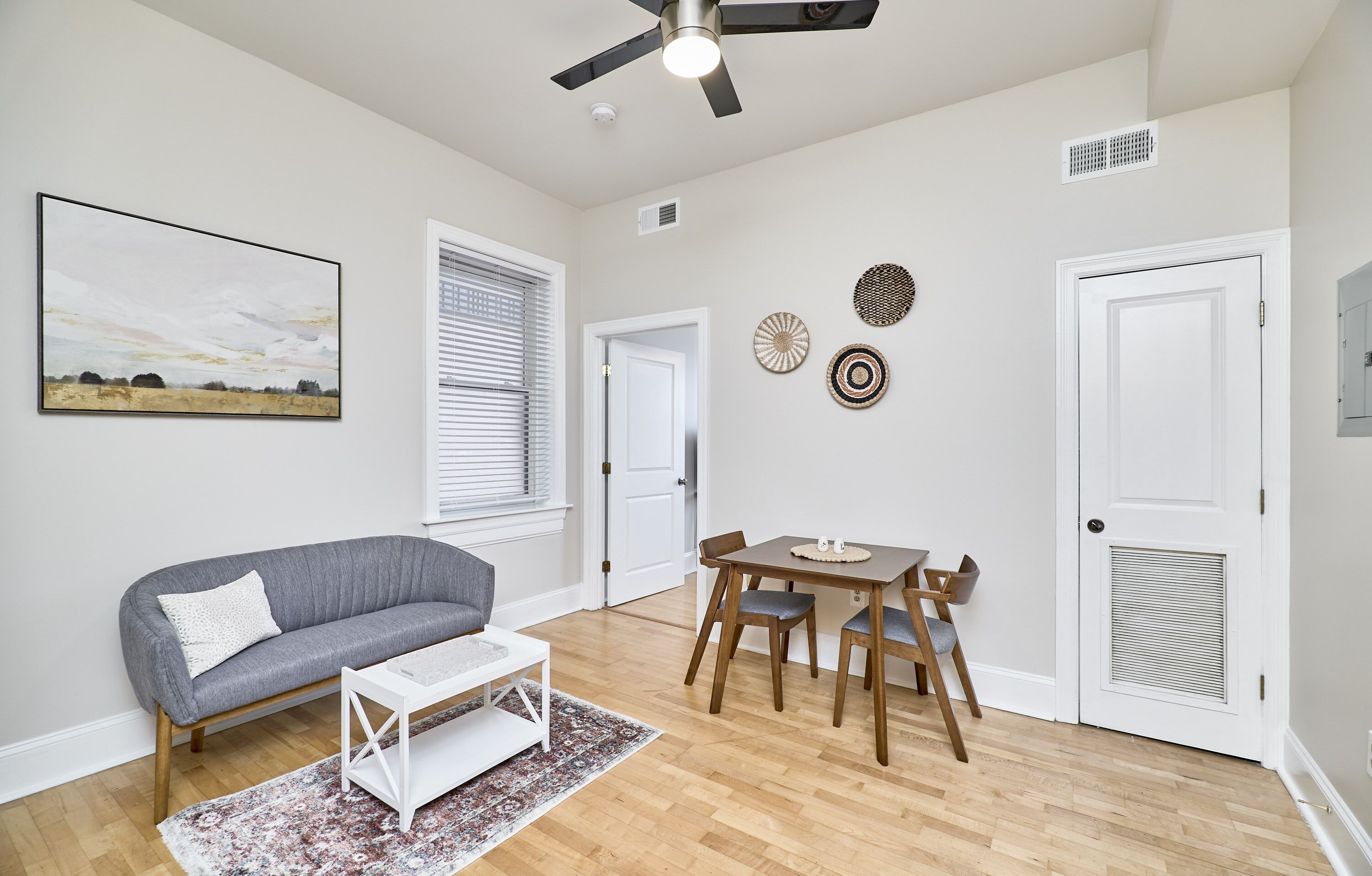 A modern living room with a gray sofa, a dining table with chairs, a ceiling fan, and a painting on the wall.