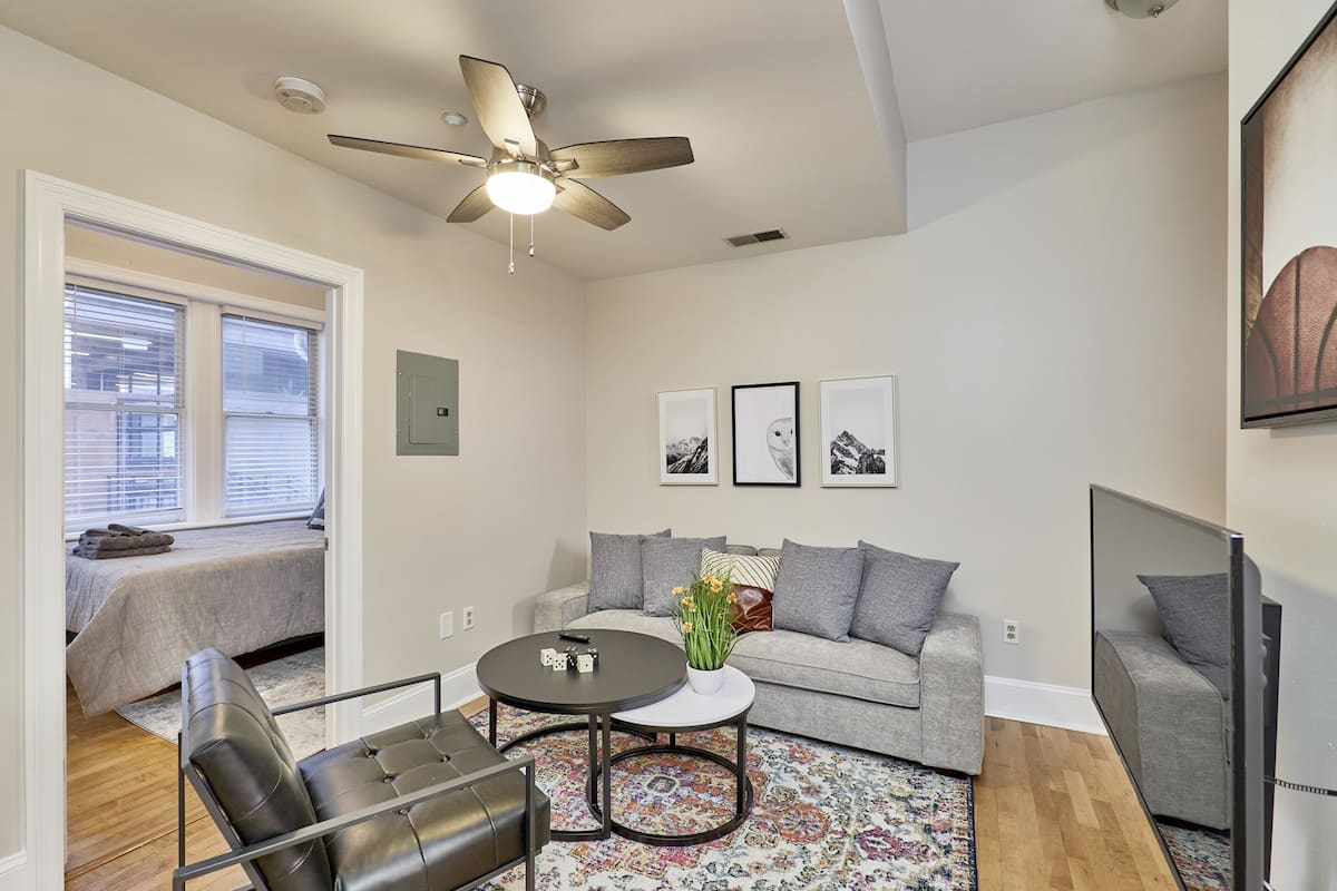 A modern living room with a grey sofa, a coffee table, a black leather chair, a ceiling fan, and a large TV.