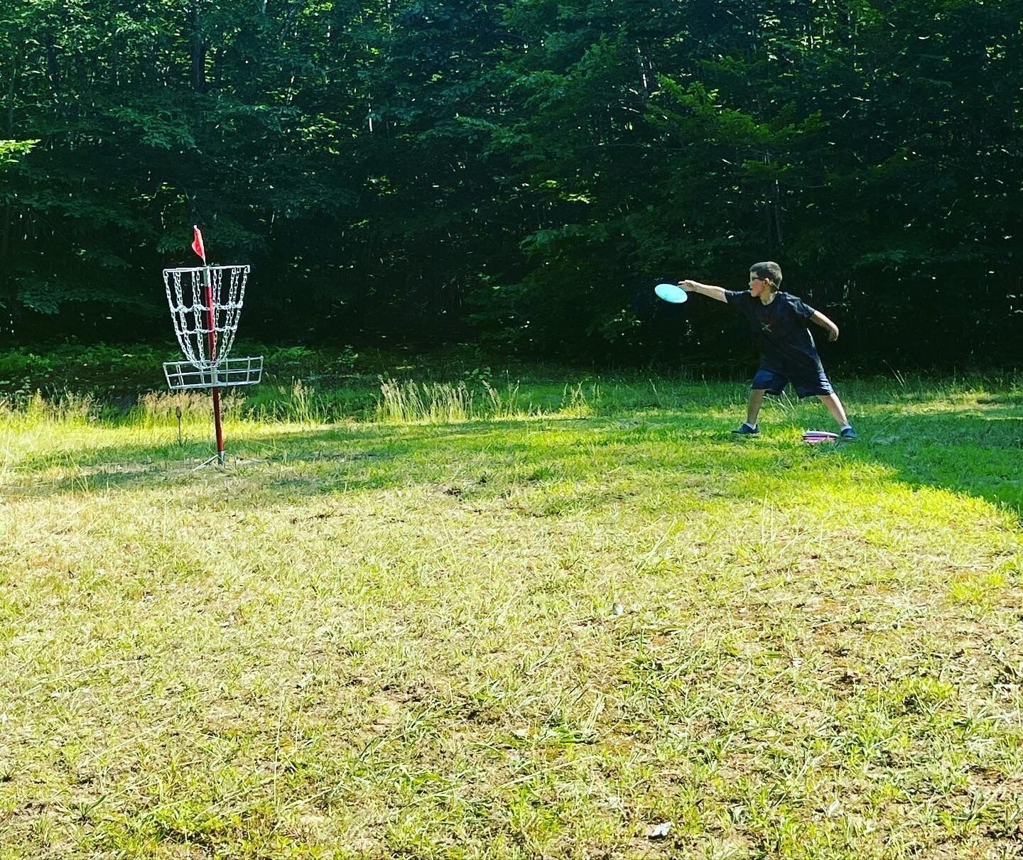 Eine Person wirft Frisbee in einer grünen Wiese mit einem Basketballkorb für Disc Golf.