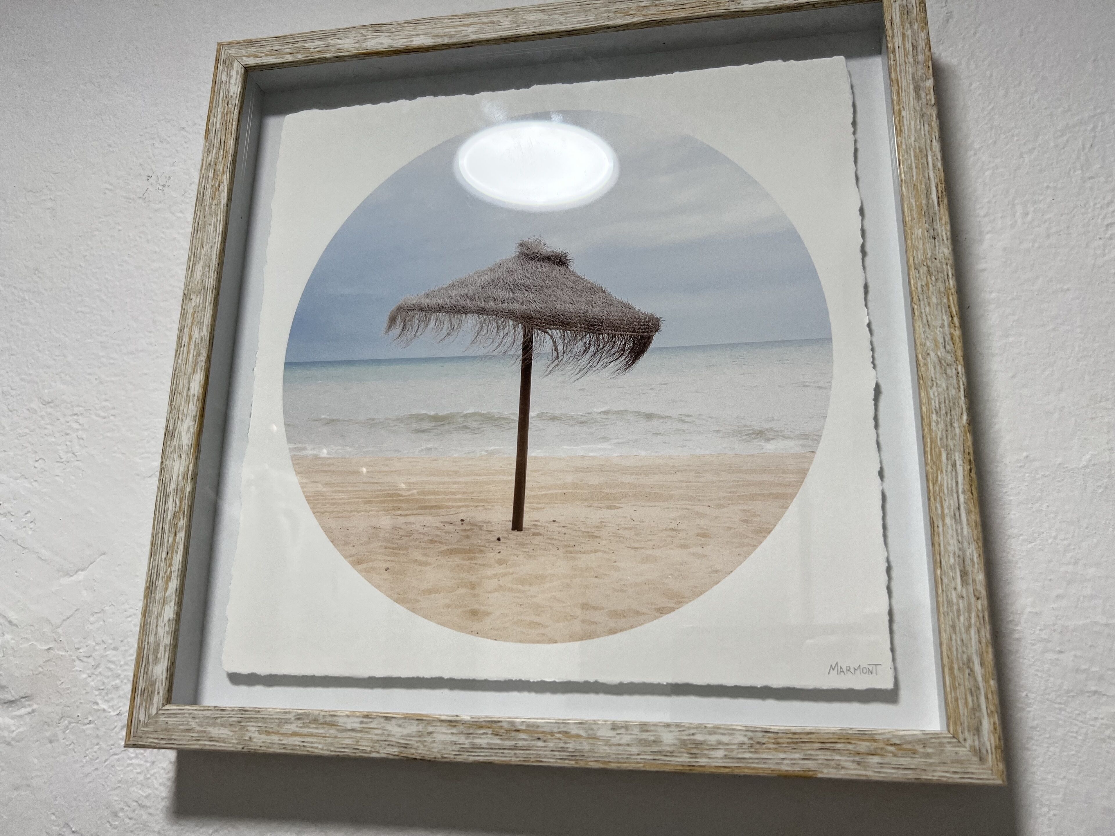 A framed photograph of a beach scene with a thatched umbrella, ocean waves, and a clear sky.