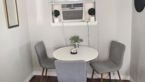 Un coin repas avec une table ronde, deux chaises grises, une plante en pot et une fenêtre avec des stores.