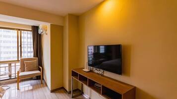 A modern hotel room with a large flat-screen TV mounted on the wall, a wooden console table, a comfortable armchair, and a large window offering a view of the cityscape.