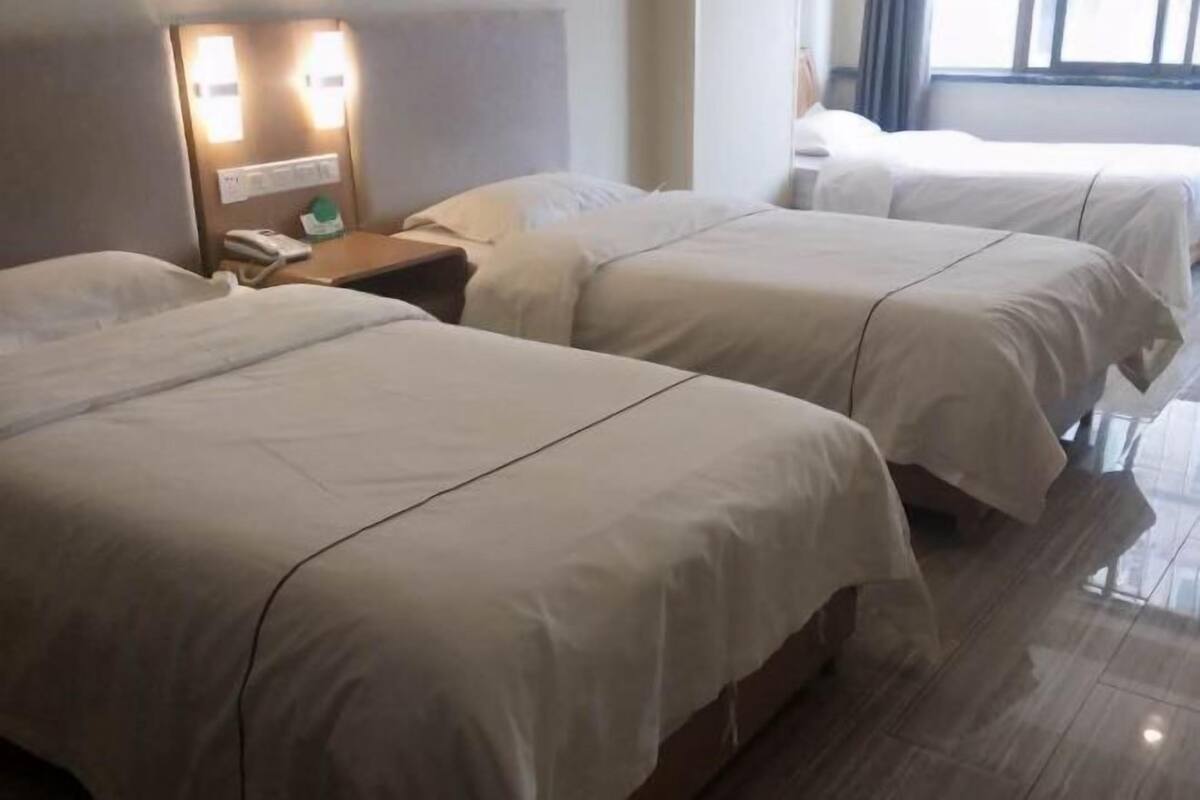 A hotel room with three beds, a bedside table with a lamp, and a window with curtains.