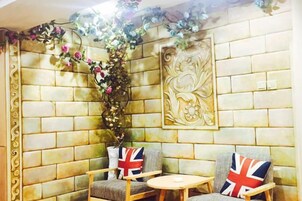 A corner seating area with two chairs, each with a British flag cushion, a small round table, and a decorative wall with a floral arrangement and a framed artwork.