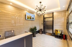 Un área de recepción moderna con pared de mármol, pantalla digital, mostrador de recepción, planta en maceta y un ascensor.