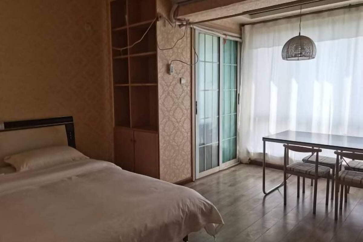 A bedroom with a bed, a wardrobe, a dining table with chairs, and a sliding glass door.