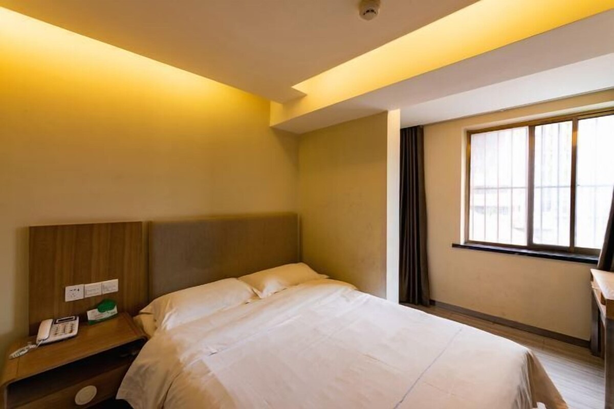 Habitación de hotel con cama, mesita de noche con teléfono y un objeto verde, ventana con cortinas y una lámpara en el techo.