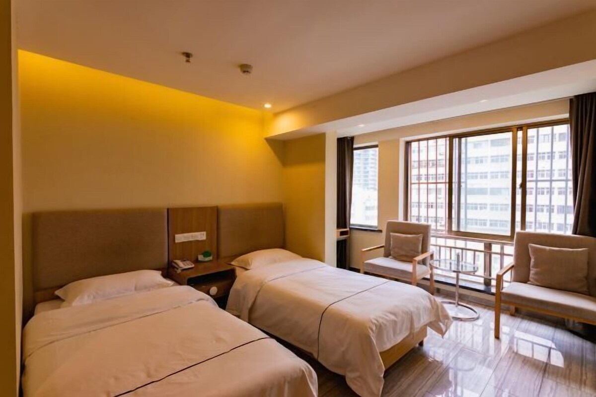 Habitación de hotel con dos camas, una zona de estar con una silla y una mesita pequeña, y un ventanal con vistas a la ciudad.