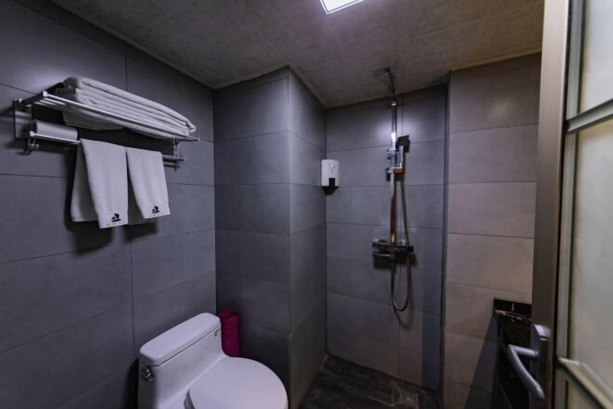 Un baño pequeño con inodoro, un perchero con toallas dobladas, ducha con cabezal de ducha manual y un cubo de basura pequeño.