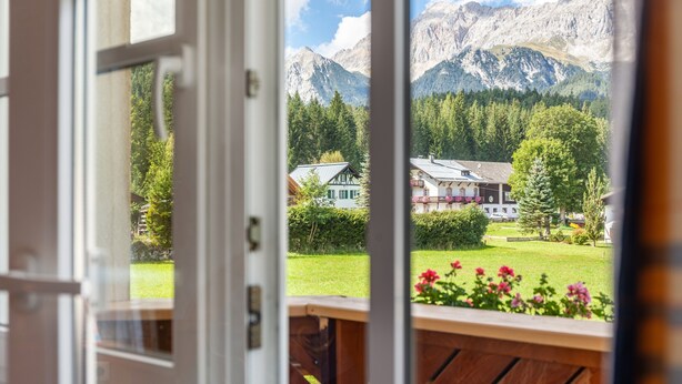 Vista a través de una ventana con un balcón de madera, un césped verde y casas al fondo con montañas.