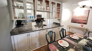 Una cocina con armarios blancos, encimera de granito y comedor con una mesa preparada para cuatro.