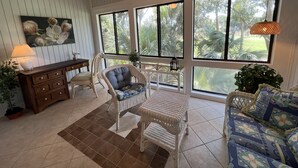 Una habitación amplia con ventilador de techo, ventanas grandes, mobiliario de mimbre y vista a zonas verdes.