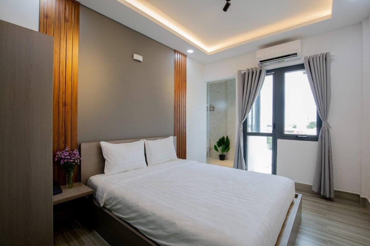 A modern bedroom with a large bed, wooden paneling, a small bedside table with a vase of flowers, and a window with curtains.